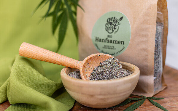 Holzschale mit gekeimten Hanfsamen und Löffel vor der Hanfland-Verpackung, daneben ein Hanfblatt